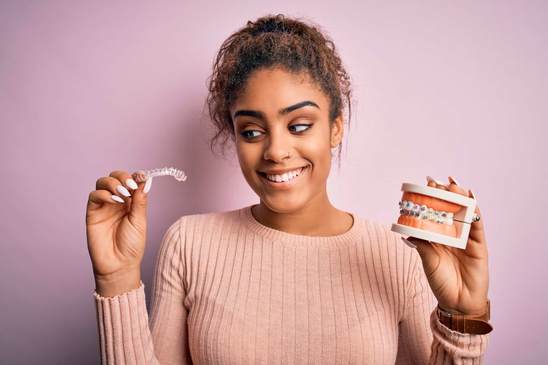 Young african american woman smiling happy holding professional orthodontic denture with metal braces and removable invisible aligner. Comparation of two dental straighten treatments